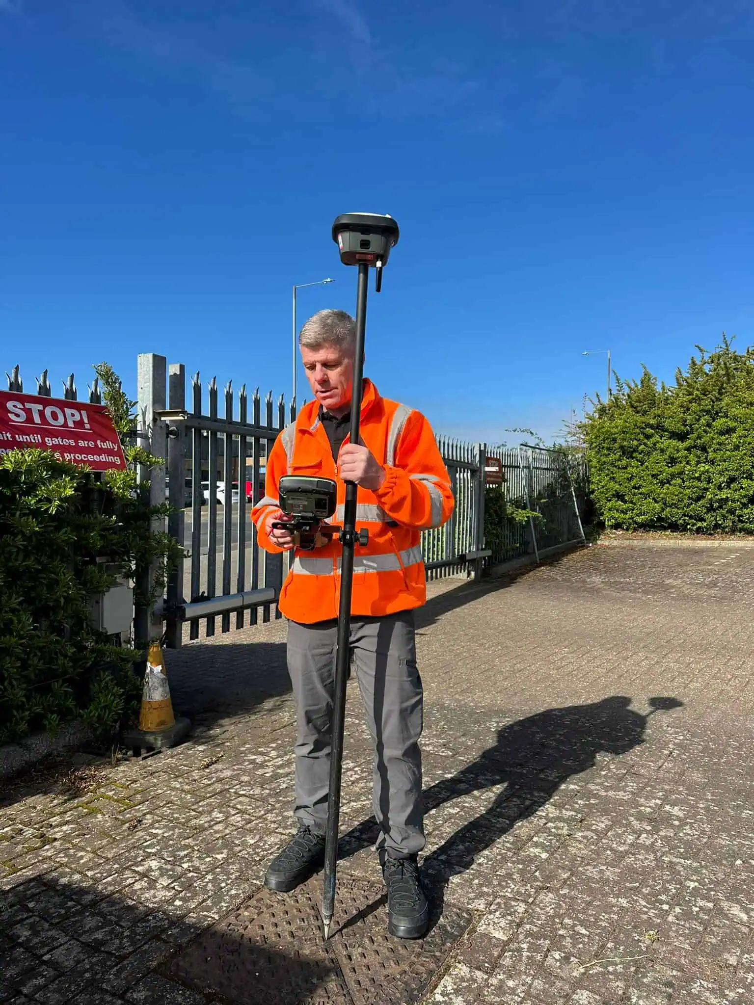 Surveyor in high-visibility jacket using a GPS rover pole and controller near a gated industrial site
